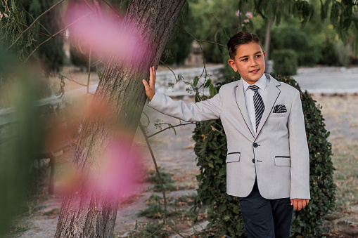 Full length of confident 8 year old schoolboy in white shirt holding tie at beige backdrop, serious look. Perfect kid boy model posing, studio shot. Learning, education concept. Copy ad text space