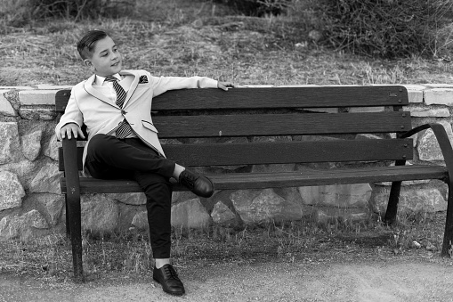 A grayscale shot of an adorable male child in a formal suit outfit posing on a bench