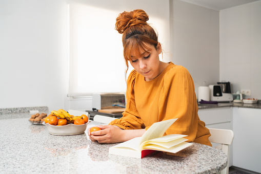 A caucasian female holding a tangerine while reading a book on a table