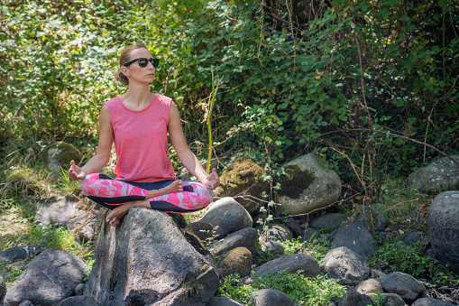 A shot of an adult Caucasian woman in a pink shirt sitting in a yoga pose on a stone in Spain
