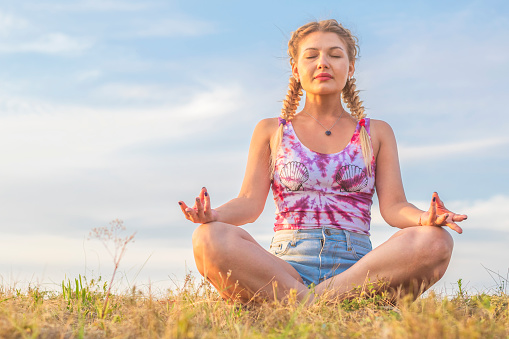 Beautiful young woman sits in a lotus position with closed eyes on a green lawn against a blue summer sky. Yoga class outdoors.