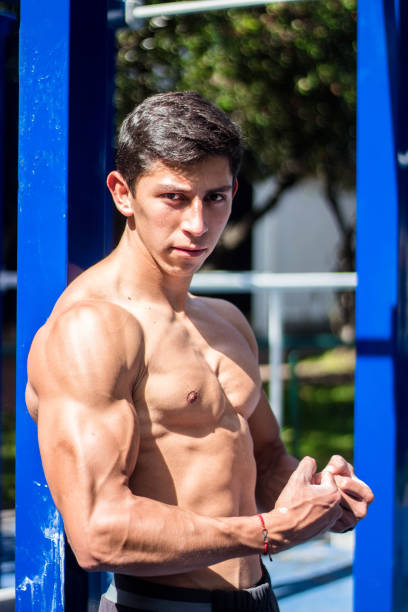 A vertical shot of a strong Hispanic bodybuilder flexing his biceps in a park
