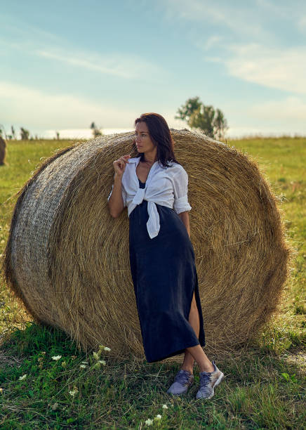 A beautiful young girl stands on a green field, leaning against a haystack. Sunny summer day. She is happy.