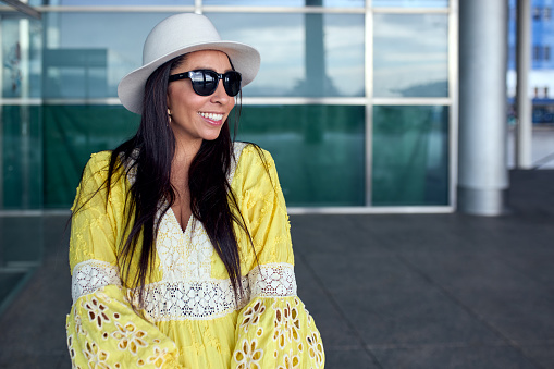 A pretty Hispanic woman in a yellow dress and dark sunglasses smiling in the street