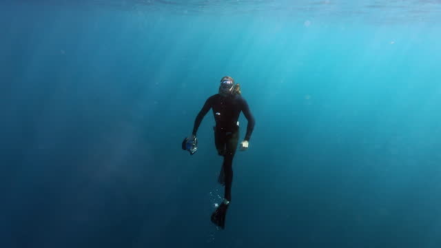 Free diving cameraman swimming to the surface from the deep blue ocean with light rays
