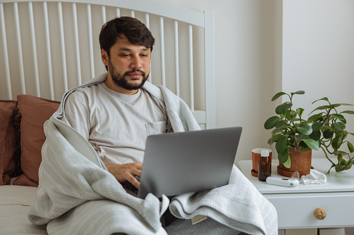 Guy with the flu with laptop works from home, he lies in cozy bed, among pillows, wrapped in a blanket