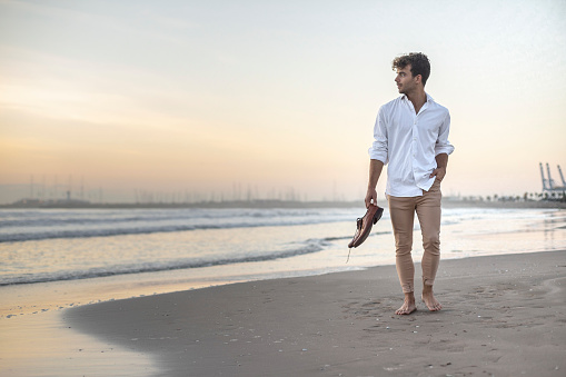 A classy elegant man in white shirt and beige pants walking on the beach with his shoes in hand