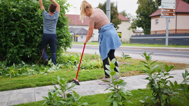 Woman brooming backyard while man is cutting plant leaves in garden