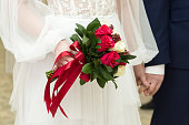 A bright bouquet with red roses in the hands of the bride on the background of a white dress