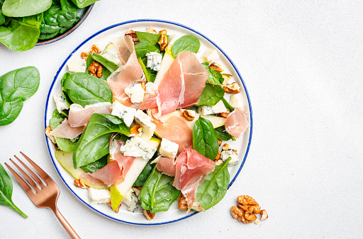 Fresh pear salad with jamon, blue cheese, spinach and nuts on white table background, top view, copy space