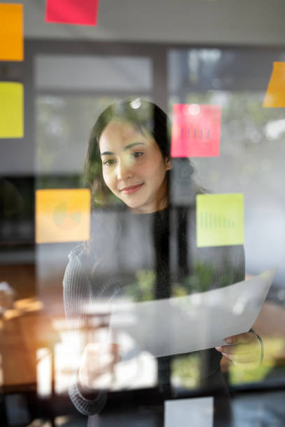 istock Young smiley attractive, businesswoman using sticky notes in glass wall to writing strategy business plan to development grow to success 1460223724