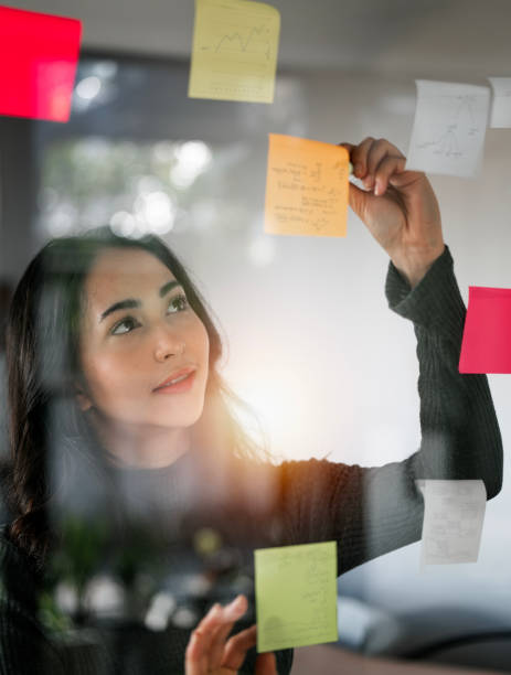 istock Young smiley attractive, businesswoman using post it notes in glass wall to writing strategy business plan to development grow to success 1460225875