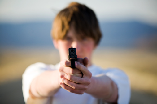 teenager pointing modern 9mm handgun at camera, shallow depth of field with focus on front of gun