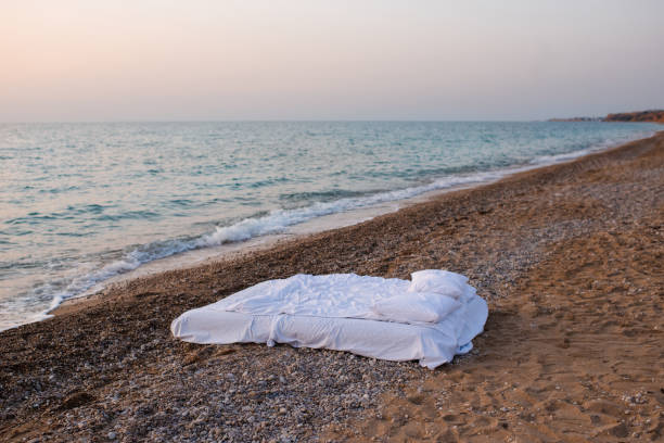 Bed with white duvet and pillows at beach with sea background over sunrise outdoor. Summer vacation season.
