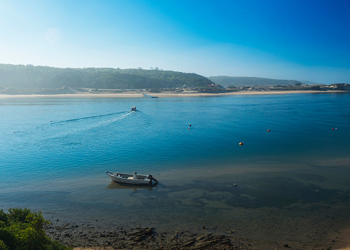 Idyllic view of the mouth of Mira river flowing into the Atlantic Ocean with anchored fishing boat and small ferry boat acrross the river. Vila Nova de Milfontes, Rota Vicentina Coast, Portugal.
