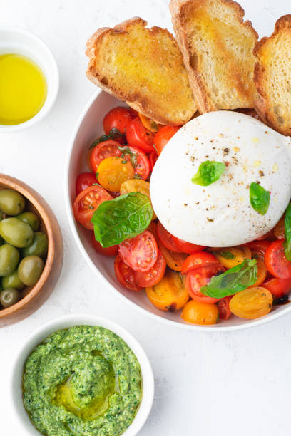 istock Farmer Italian soft cheese mozzarella di bufala campana served with fresh basil and red and yellow cherry tomatoes, croutons, homemade pesto, olives. Healthy Italian summer food, white table, close-up 1460238172