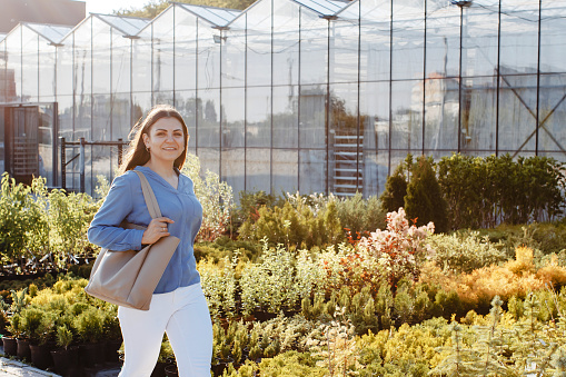A young pretty woman walks down the street near the large greenhouses. A woman goes to buy green plants for decoration. Landscaping concept