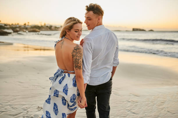 praia, tatuagem e amor com um casal de mãos dadas enquanto caminham juntos na areia à beira-mar. de volta, viagem e verão com um homem e uma mulher se unindo no mar para um passeio romântico na costa - tattoo women back rear view - fotografias e filmes do acervo