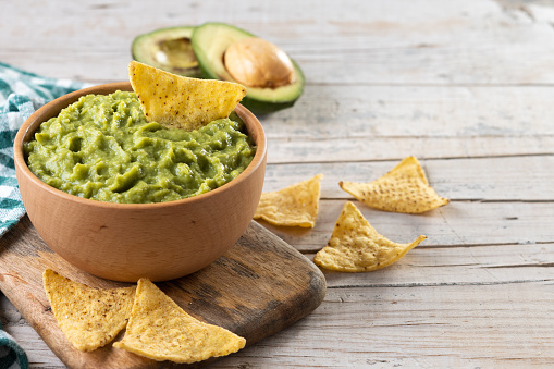 Mexican food: guacamole dipping sauce in a bowl with tortilla chips shot from above on abstract bluish tint table. Ingredients for preparing guacamole are all around the bowl and includes ripe avocados, tomatoes, lime, cilantro and salt. Predominant colors are green and blue. High resolution 42Mp studio digital capture taken with Sony A7rII and Sony FE 90mm f2.8 macro G OSS lens