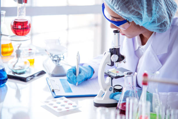 istock Woman scientist hand write note information in medical lab look at science microscope medical test, research biology chemistry. Asian Females technician laboratory analyzing blood test pharmaceutical 1460248583