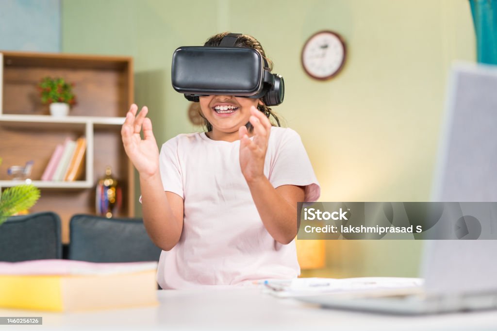 happy smiling teenager Girl kid with vr or virtual head set experiencing online or virtual class using hand gesture in front of study table at home - concept of cyberspace, technology and entertainment. Asian and Indian Ethnicities Stock Photo