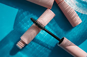 pink clean makeup brush for mascara lies on the open tube, near to closed tubes of cosmetics, lip gloss, liquid lipstick, pink eyeliner, glass vase on a blue background with shadows. Copy space