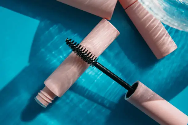 Photo of pink clean makeup brush for mascara lies on the open tube, near to closed tubes of cosmetics, lip gloss, liquid lipstick, pink eyeliner, glass vase on a blue background with shadows. Copy space