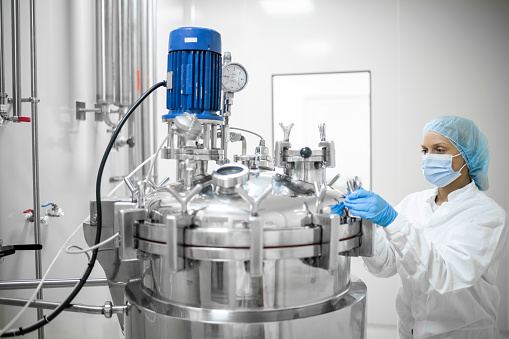 A young female skilled worker in protective gear operates a high-tech equipment in a pharmaceutical production facility, ensuring the precise mixing of substances to produce life-saving medications. Highlighting the dedication and precision required in the industry.