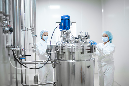 Two young female workers in protective gear operate a stainless steel tank and vessel in a pharmaceutical production facility. The tank is equipped with an agitator for precise mixing of substances, ensuring the smooth production of life-saving medications. The facility employs advanced technology and strict controls to maintain the sterility and efficacy of the drugs being produced.