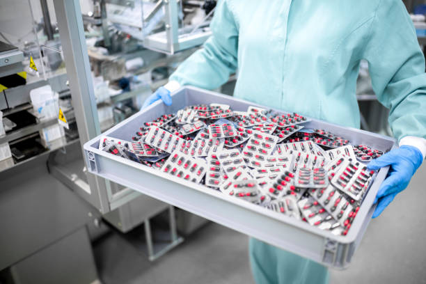 A worker in protective gear carefully carries a container filled with blister-packaged capsules stock photo