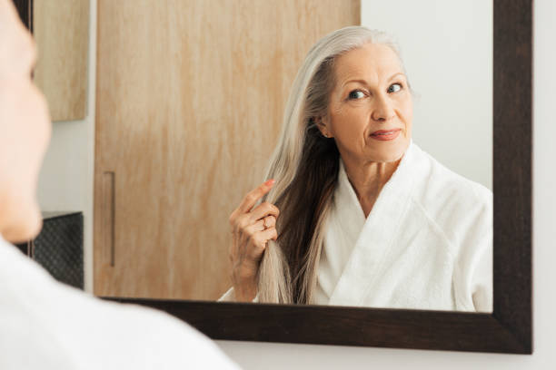 Senior woman examine her long grey hair while standing in front of a bathroom mirror stock photo