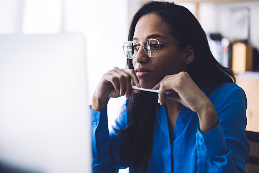 Black creative thoughtful smart concentrated lady in eyeglasses thinking about strategy sitting on computer in workplace and leaning with chin on pen