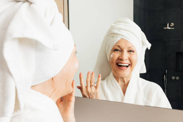 смеющаяся престарелая женщина с завернутым полотенцем на голове стоит перед зеркалом в ванной комнате - st petersburg стоковые фото и изображения