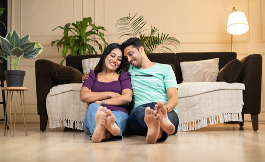 wide shot of happy smiling indian couple embracing each other by eyes closed at home while sitting on floor - concept caring husband, newly married couples and relationship bonding.