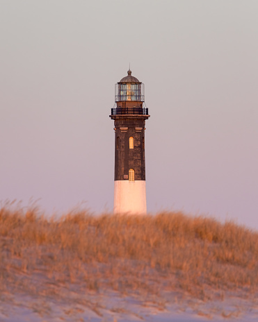 Tall stone lighthouse seen over sand dunes and beach grass with a soft pink sky. Fire Island, Long Island New York