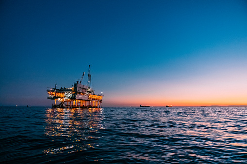A striking image of an offshore oil rig at sunset off the coast of Huntington Beach, California. The pink & orange tones of the setting sun highlight the industrial machinery and equipment used in the drilling and extraction of fossil fuels, including crude oil and natural gas. 

The scene captures the intersection of the energy industry and the beauty of the Pacific Ocean. The image speaks to issues of fuel and power generation, energy crises, and environmental concerns surrounding the oil and gas industry.