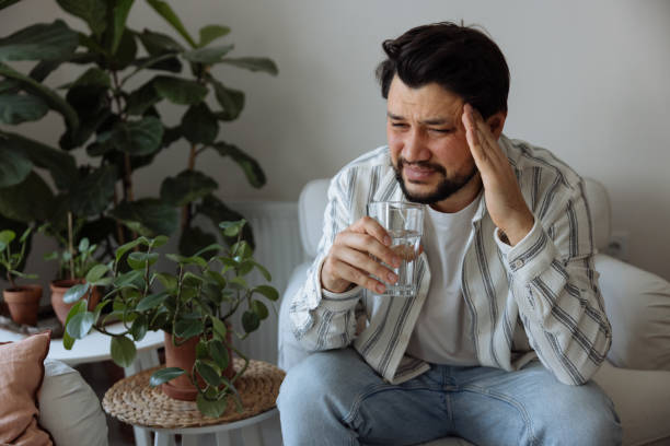 Young man suffering from headache, migraine or hangover at home Young man suffering from strong headache or migraine sitting with glass of water in the kitchen, millennial guy feeling intoxication and pain touching aching head, morning after hangover concept hangover stock pictures, royalty-free photos & images