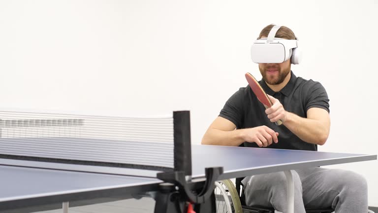 An adult man with a disability wearing virtual reality glasses plays table tennis in a bright hall.