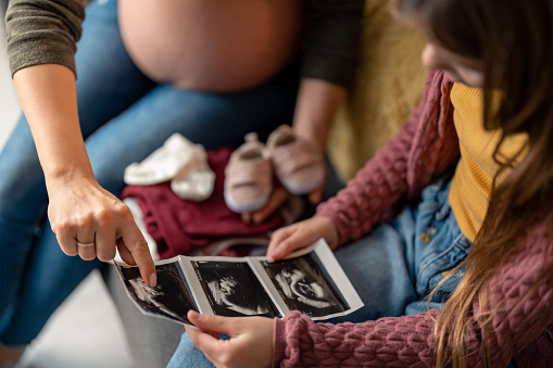 Cropped shot of unrecognizable pregnant woman sitting next to her daughter pointing on ultrasound picture, showing, explaining to her how the baby looks right now, holding pair of non focused pale pink shoes.