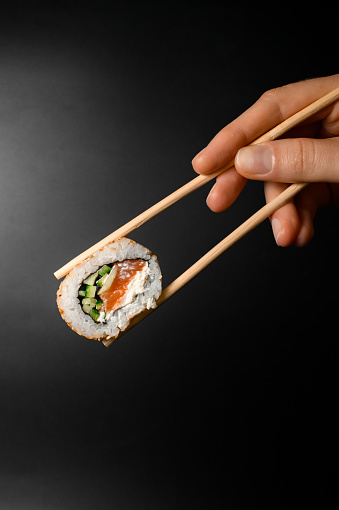 hand holding one piece of roll california with smoked salmon, cream cheese, cucumber garnished sesame seeds with bamboo chopsticks on dark background.