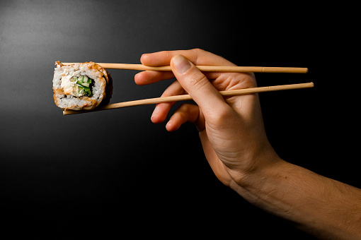 female hand skilfully holds Philadelphia sushi roll with smoked eel, cucumber, avocado, cream cheese with bamboo chopsticks on dark background. Japanese food.