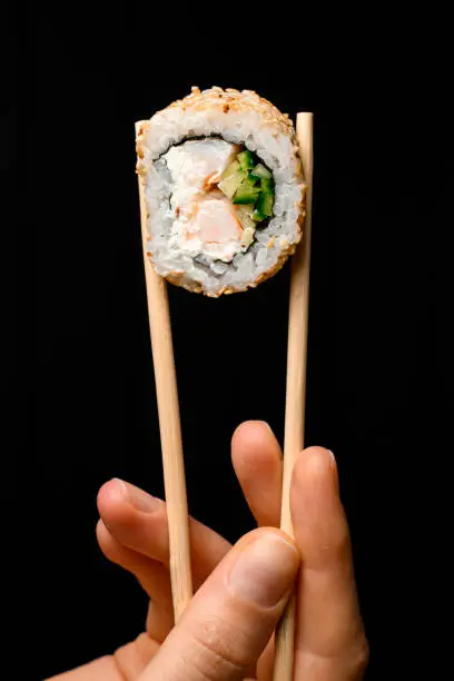 Close-up view of one piece of roll california with snow crab meat, cream cheese, cucumber garnished sesame seeds in bamboo chopsticks in hand on dark background.
