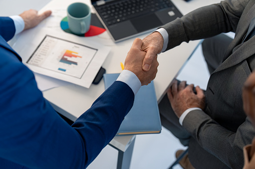 Business people shaking hands. Recruitment concept to hiring of a new talented specialists for international company. Paper documents containing graphs and financial data, laptop, notebook and cup on table.