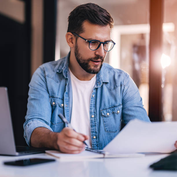 istock Satisfied young man with glasses sitting at a desk and doing paperwork at his workplace. 1460311368