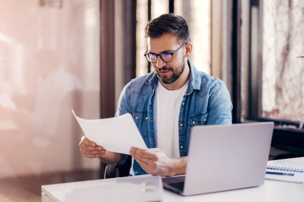 istock Satisfied young man with glasses sitting at a desk and doing paperwork at his workplace. 1460311907