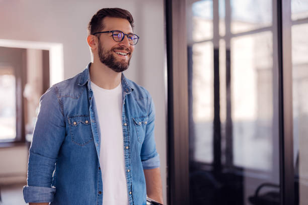 Cheerful entrepreneur in denim shirt, wearing glasses, standing in his office in the morning. stock photo