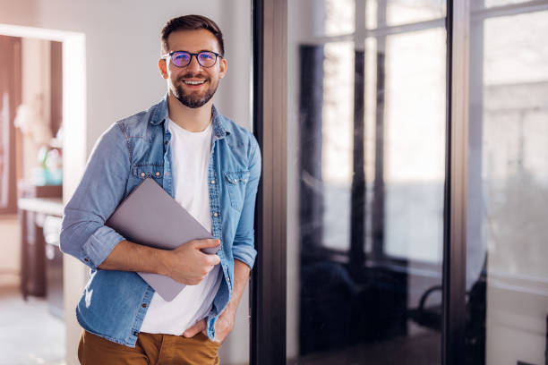 retrato de un joven hombre de negocios moderno de pie sosteniendo una computadora portátil y mirando a la cámara con una sonrisa feliz - only men mid adult men men author fotografías e imágenes de stock