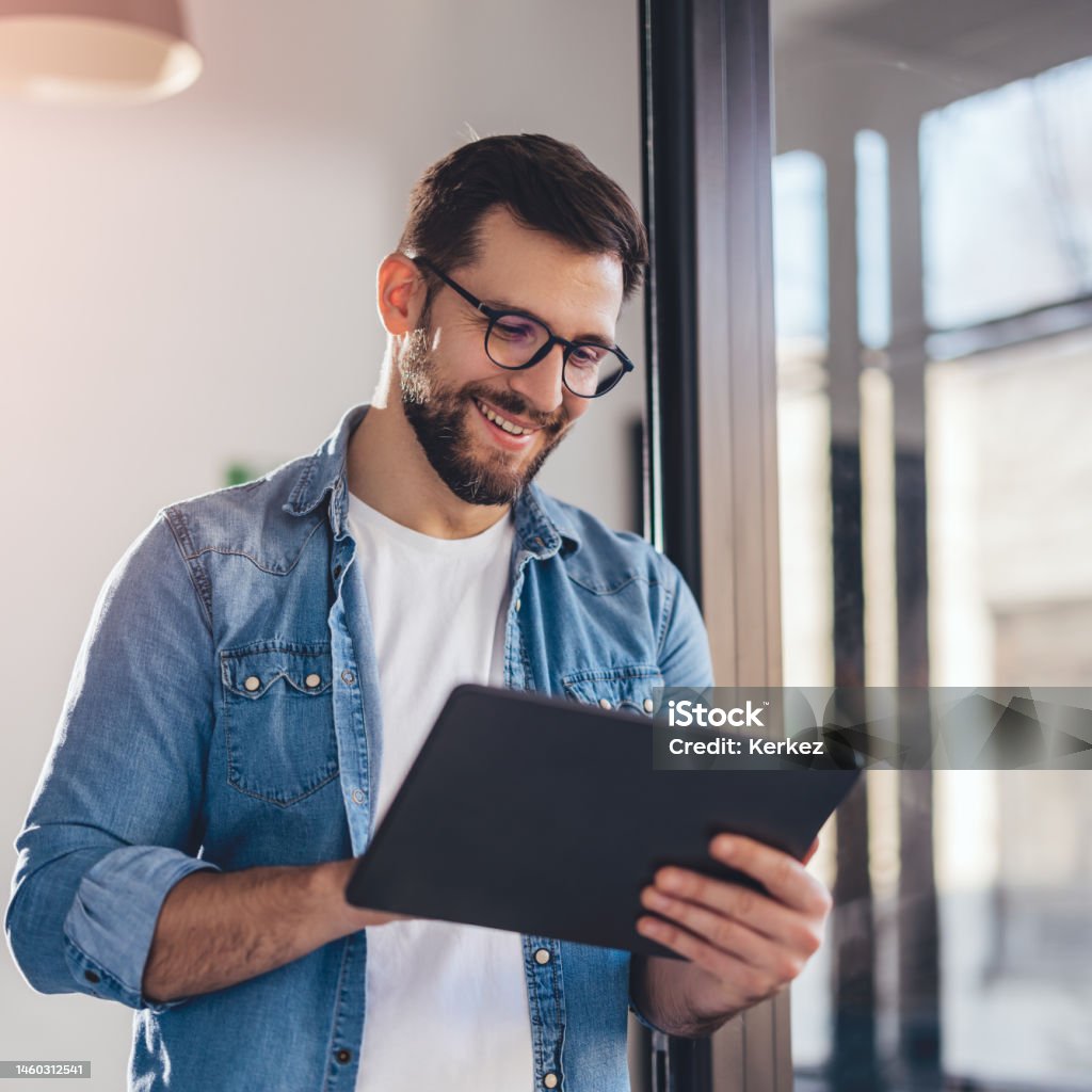 Smiling young businessman working online with digital tablet while standing by window. Men Stock Photo