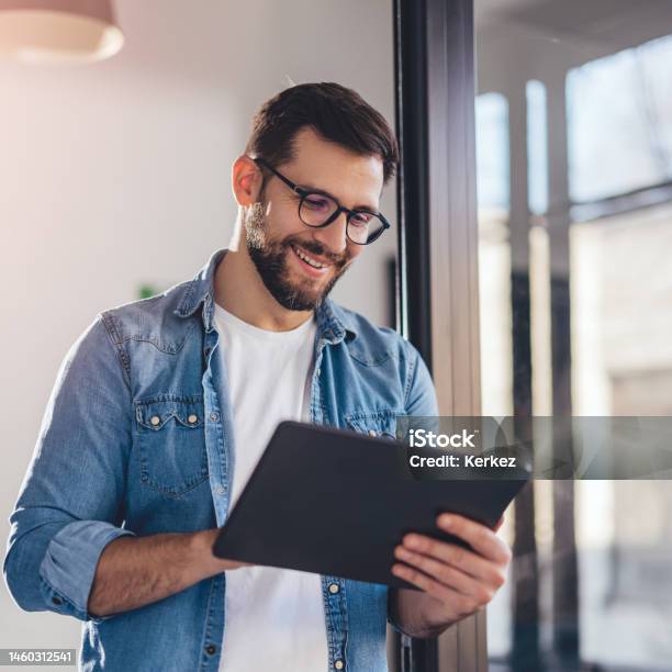 Smiling Young Businessman Working Online With Digital Tablet While Standing By Window Stock Photo - Download Image Now