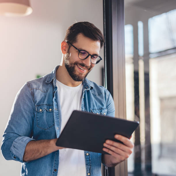 smiling young businessman working online with digital tablet while standing by window. - surfplatta bildbanksfoton och bilder